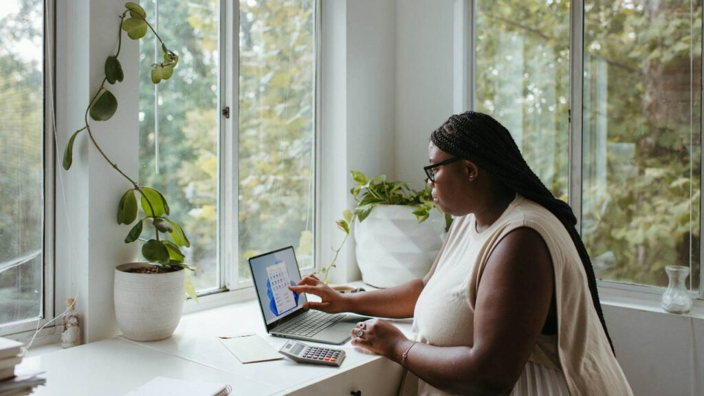 A woman using a laptop and a calculator in a room with large windows and plants