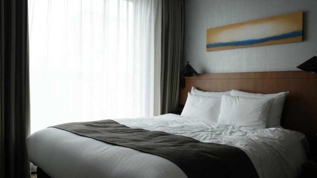 A photo of a plush hotel bed located next to a window with a curtain 